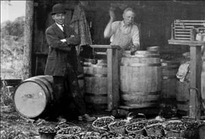 A smiling man in a bowler hat, suit and tie next to a man hammering a barrel in a barn around buckets of produce
