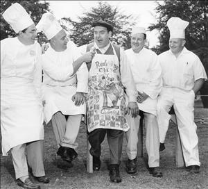 Rosellini between 4 cooks in chef's whites one of whom is feeding him a hot dog, while wearing a decorated apron with an illustration of a man on a scale and the word "Calories" visible