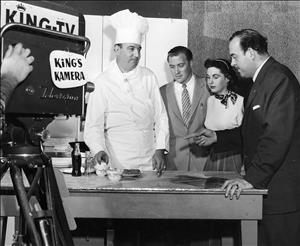 Rosellini standing beside two people and a cook on a movie set with camera in the foreground labeled "King TV, King's Camera"