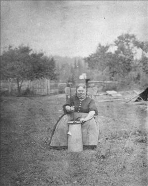 Fares sitting on chair in field, churn in front on ground, right hand holding churn handle wearing dark blouse with buttons, wide checkered apron. Trees and fence in background.