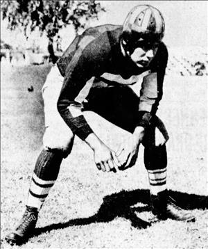 Harris crouched in a football uniform with striped socks and early, leather helmet with a tree in the distance