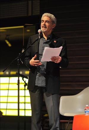Gayton speaking into a microphone on stage surrounded by molded chairs with papers in his hand