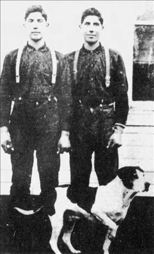Two young men with dark hair wearing suspenders and work clothes posing in front of a shingled house and window and behind a dog that is pointing with leg raised