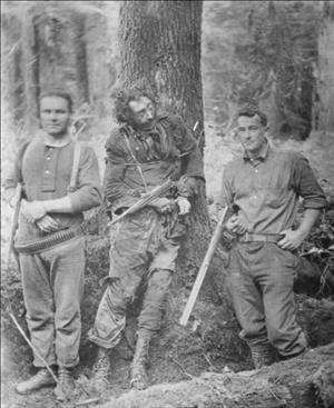 Tornow, deceased, propped up against a tree with hands bound and a rifle stuck between his arms, flanked by two men posing with rifles