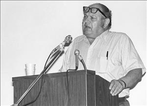 Hugo in a striped button up short sleeved shirt with a pen in his chest pocket and glasses resting on his forehead as he speaks into two microphones behind a podium