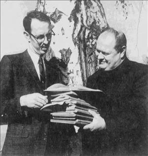 Hugo in a dark turtleneck and suit jacket in front of a tree smiling as he holds a tall stack of notebooks over to a man squinting down at the books, wearing horn rim glasses, suit and tie