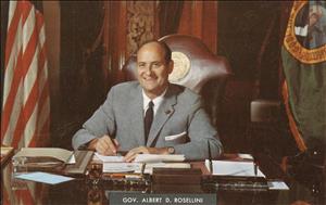 Rosellini smiling, seated in a leather chair with the Washington State emblem embedded in gold, behind a desk holding a pen to a notepad with the American and Washington State flag on either side of him