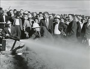 Rosellini in a suit and hardhat, leaning over to release a valve from a pipe that is powerfully spraying liquid as a crowd of journalists and spectators watch