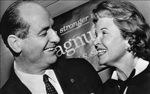 A canted angle photo of Rosellini and his wife smiling closely at one another in front of a sign reading "stronger" "Magnuson" "U.S. Senate" 