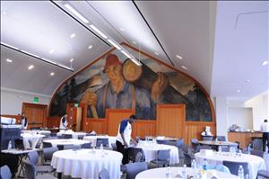 Food service workers arranging items on dining tables in a hall in front of a large mural of a lumberjack in a red hat holding timbre and an ax