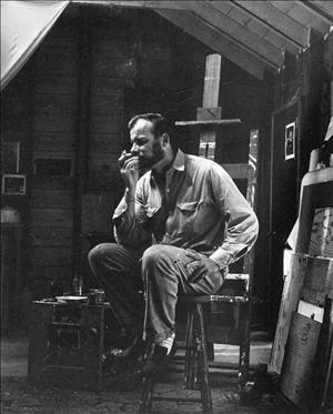A younger Meitzler smoking with a short cropped beard on a stool in front of an easel in a wood paneled studio streaked with natural light
