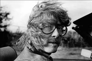 Robbins in thick, round glasses and mustache riding in a convertible with windswept hair