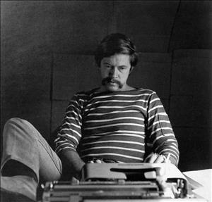 Robbins in a striped shirt looking at a typewriter with his leg resting on desk