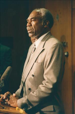 An older Charles V. Johnson in a suit speaking behind a podium