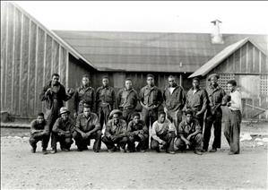 Two rows of black men, standing and kneeling in front of a tar paper structure. They are wearing work clothes. Two men, one on either end of the group, are holding up boxing gloves. 