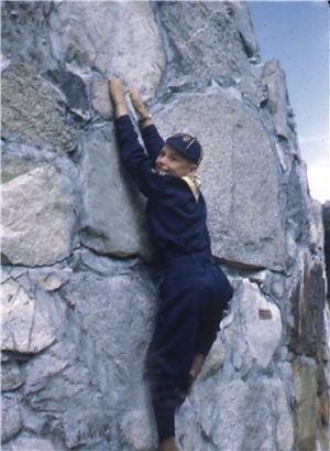 A child in a blue scouts uniform clings to a rock while climbing a rock wall