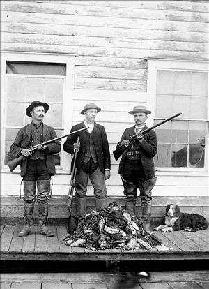 Three white men in hats and suits hold rifles while standing on a porch with a dog and a pile of dead ducks at their feet