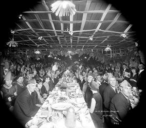 A vignette view of the interior of a dining hall filled with attendees in evening wear, wearing party hats in front of empty plates, underneath festive decorations hung from the ceiling, with text to the bottom right reading "15th Annual Banquet, Alpine, Photo Ticket 3152" 