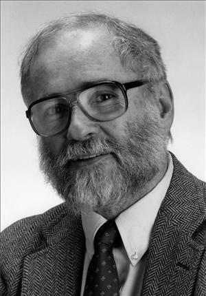 A head and shoulders portrait of a smiling white man with a beard wearing a tie and tweed coat. 