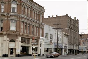 A small town street with one parked car. Brick buildings that are two and three stories high line the street. A sign outside on of the storefronts says Tammy's Tavern. 