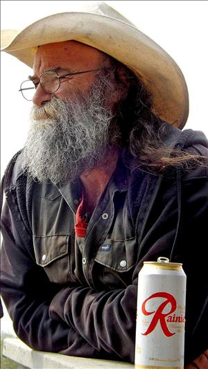 An older white man with a long gray beard and round eyeglasses, wearing a floppy cowboy hat and a weathered work shirt, leans on a railing with his arms folded. A can of beer is next to him on the railing. 