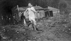 Two people in tank tops and shorts run down a dirt path. Behind them onlookers clap and cheer. There is a small one story wooden cabin on the edge of the forest in the background.