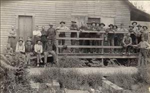 A long row of workers standing and sitting on the deck of a one story wooden building with dirt and shrubs in the foreground.