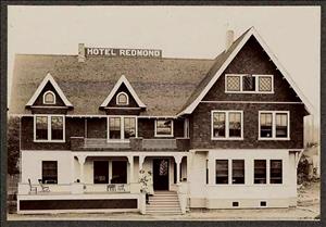 A large two story building with sharply peaked roofs, two chimneys, and a long front porch. The sign on the roof says Hotel Redmond. 