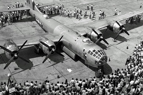 Elevated view of the unveiling of Boeing B-29 Superfortress bomber, sitting on tarmac surrounded by crowds of people looking at its four propeller engines, armaments, cockpit windows, and other features.  