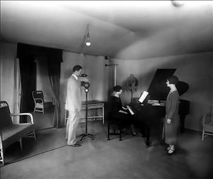 A woman sits at a piano while another woman in a dress stands beside the piano and a man stands in front of a microphone. 