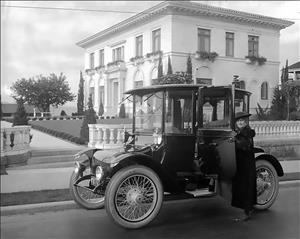 A woman in a dress coat and hat stands by the open door of an early twentieth century automobile. A two story white mansion in the background. 