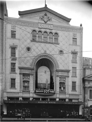 A tall stone building with arched windows and an arched marquis. A sign on the ground floor awning says Rhodes Brothers and the word Rhodes is carved into the top of the building.