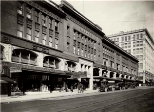 A brick building on a city street with several storefronts. Automobiles are parked in front. 