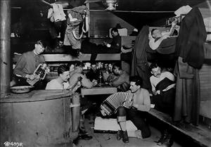 Inside a small room men engage in various activities. Some are in bunks beds reading books. One stands with an oil lantern. Two are seated with a guitar and accordion. Several sit around a table playing cards. 