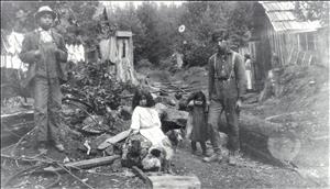 Children and young adults wearing work cloths and dresses beside a dog and in front of lumber and wood panel cabins