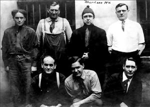 Seven men pose for the camera in front of jail cells. Three are sitting. Four others stand behind them. 