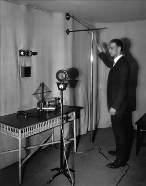 A man in a small room taps a tall metal pole. In front of him is a microphone on a stand and a small table with tools on it. 