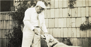 A medium sized dog holds a newspaper in its mouth for a man in a cap standing in front of a house with cedar shingles. 