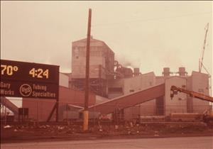 Industrial buildings partially obscured by red dust and smog. A large digital clock in front of the buildings says it is seventy degrees at four twenty four P.M.