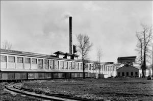 Railroad tracks lead up to a long factory building with a smokestack belching smoke. 