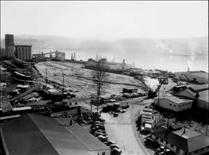 A road with many cars parked along it leads up to several industrial buildings and silos on the banks of a large river. 