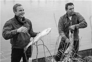 Two men, one white and one indigenous, stand in a boat on the river's edge holding large fish