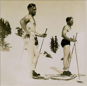 Two men in nothing but short shorts and boots standing on skis and holding ski poles on a snowy slope. 