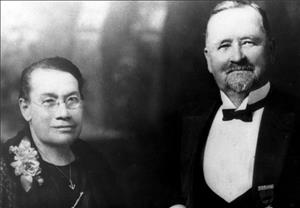 A head and shoulders portrait of a smiling couple. On the left, a woman with eyeglasses, a flower corsage, and an anchor necklace. On the right, a man in a tuxedo. 