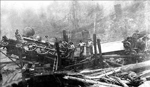 Several men stand on and around a wrecked locomotive that is crumpled between logs and debris. 