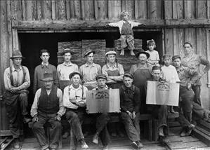 A group of men in work clothes and hats sit and stand for the camera, two of them holding large shingles. There are three children in the picture, one of whom stands on a stack of wood with arms spread wide. 