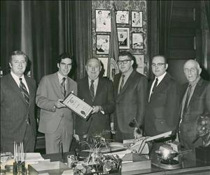 Six white men in suits stand behind a large desk smiling. Two of them are holding a framed certificate. The image is black and white. 