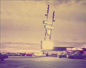 A mid century hotel sign glows in pink evening light above a parking lot full of cars from the 60s. The sign says Ocean Shores Inn. A one-story building at the end of the parking lot. 