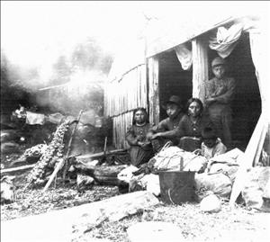 Five indigenous adults and children sit outside a wooden structure with bushels stacked in front of them. 