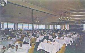 A room full of tables with white tablecloths and gold colored dining chairs. Mid century chandeliers hang from the ceiling and the ocean is visible through multiple windows along the far wall. 